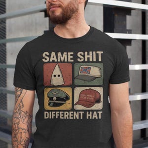 May include: Dark gray t-shirt with the text "SAME SHIT DIFFERENT HAT." The shirt features four images: a white hood, a Confederate flag hat, a military hat, and a red "Make America Great Again" hat.