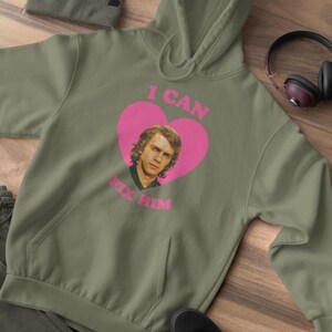 May include: Olive green hoodie with a pink heart graphic featuring a portrait and the text "I CAN FIX HIM." The hoodie has a kangaroo pocket and a drawstring hood. A matching beanie, pants, and black shoes are also visible.
