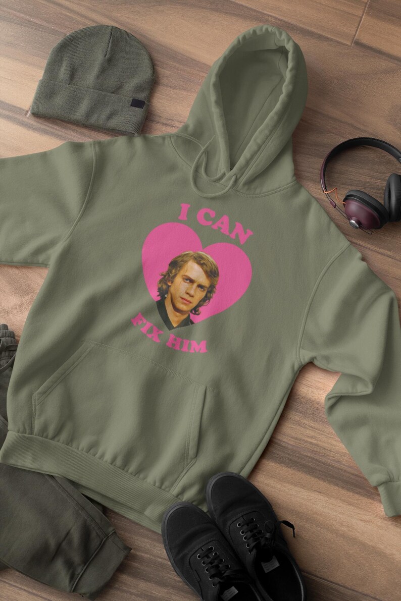 May include: Olive green hoodie with a pink heart graphic featuring a portrait and the text "I CAN FIX HIM." The hoodie has a kangaroo pocket and a drawstring hood. A matching beanie, pants, and black shoes are also visible.