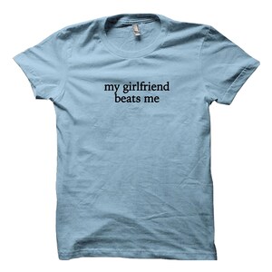 May include: Light blue t-shirt with the text "my girlfriend beats me" printed in black. The shirt has a crew neck and short sleeves. The fabric appears to be a soft cotton blend.
