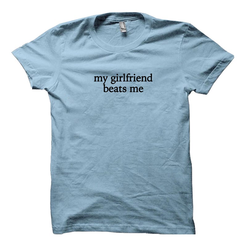 May include: Light blue t-shirt with the text "my girlfriend beats me" printed in black. The shirt has a crew neck and short sleeves. The fabric appears to be a soft cotton blend.