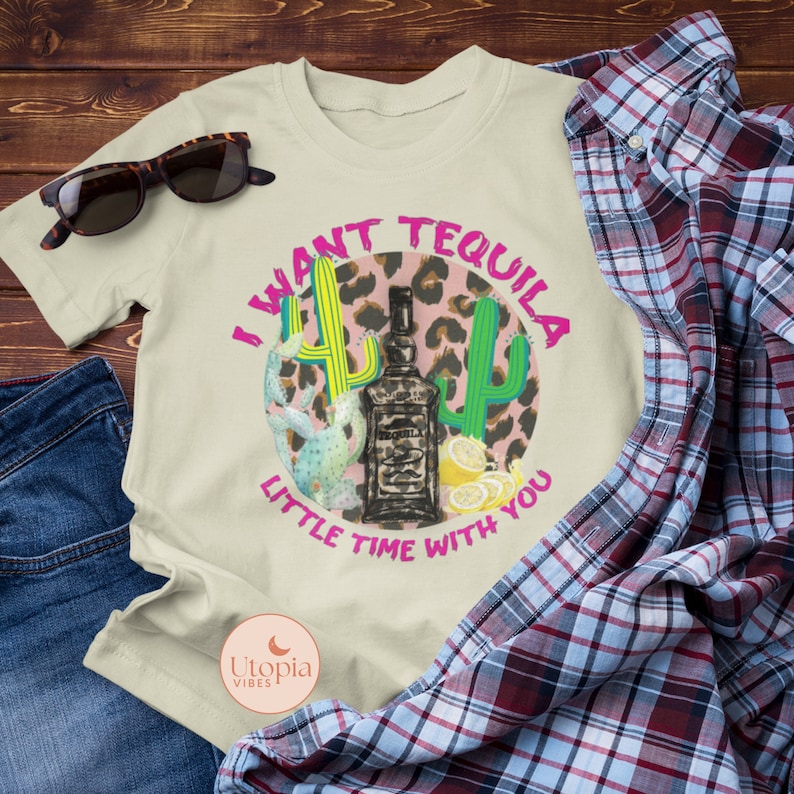 以下が含まれることがあります： ピンク、グリーン、ブラックのグラフィックデザインが施されたベージュのTシャツ。グラフィックには、テキーラのボトル、レモン、サボテンが描かれています。グラフィックのテキストは「I want tequila little time with you」です。