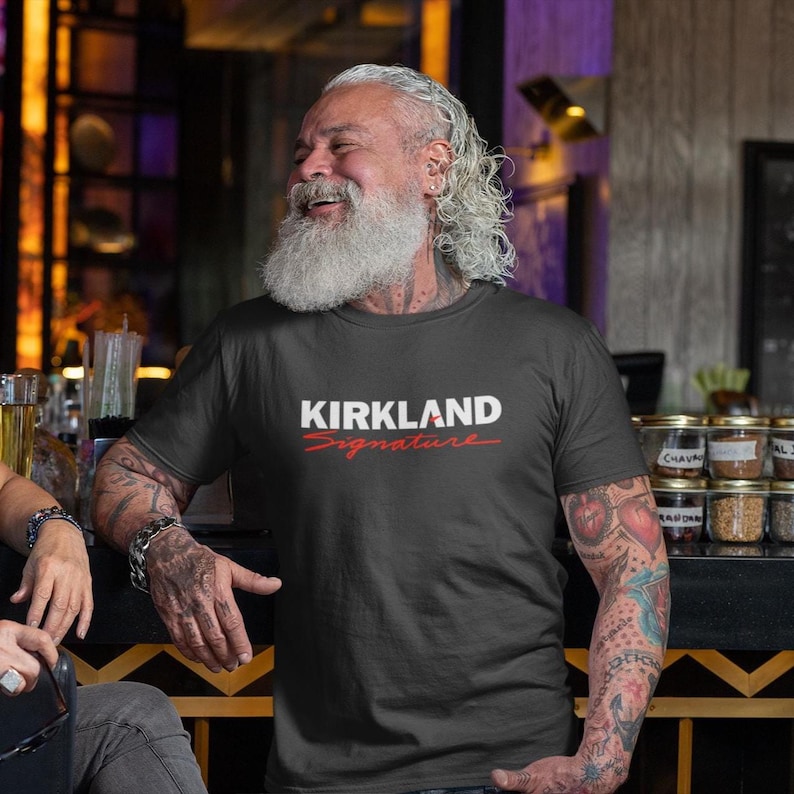 Puede incluir: Camiseta negra con el texto "KIRKLAND Signature" en blanco y rojo. El hombre sonr&iacute;e y se apoya en una barra. El hombre tiene una larga barba blanca y tatuajes en los brazos.