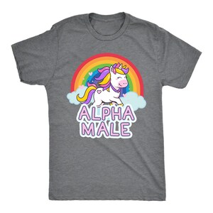 May include: A heather gray t-shirt featuring a cartoon unicorn with a rainbow and the text "ALPHA MALE" in pink. The unicorn has a pink nose, yellow horn, and purple and yellow mane. The shirt is a crew neck.