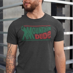 Może przedstawiać: Szary t-shirt z nadrukiem w kolorze zielonym i czerwonym z napisem "Mountain Dude". Grafika przedstawia stylizowaną sylwetkę osoby jeżdżącej na nartach.