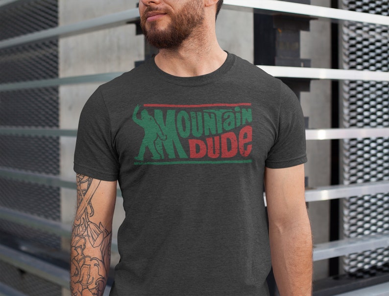 Może przedstawiać: Szary t-shirt z nadrukiem w kolorze zielonym i czerwonym z napisem "Mountain Dude". Grafika przedstawia stylizowaną sylwetkę osoby jeżdżącej na nartach.