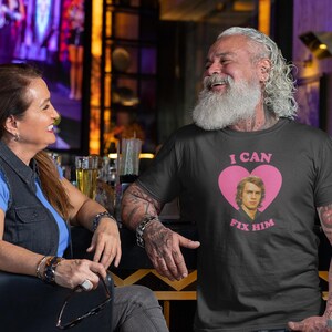 May include: A dark gray t-shirt with the text "I CAN FIX HIM" in pink, along with a pink heart and a portrait of a man. The shirt is worn by a person with a long white beard and tattoos.