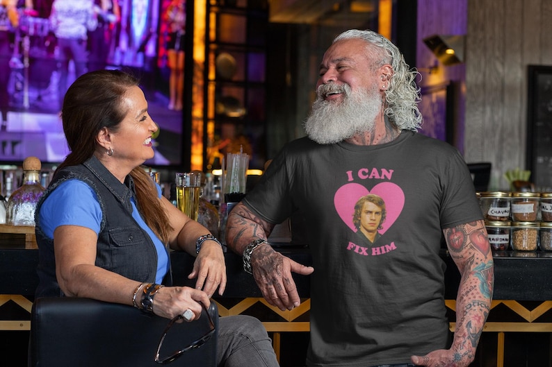 May include: A dark gray t-shirt with the text "I CAN FIX HIM" in pink, along with a pink heart and a portrait of a man. The shirt is worn by a person with a long white beard and tattoos.