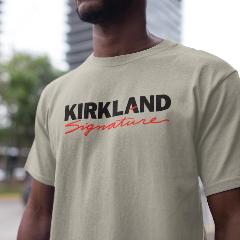 Puede incluir: Camiseta beige con la inscripci&oacute;n "KIRKLAND Signature" en negro y rojo. La camiseta de manga corta es de material suave. El texto es en negrita, sin serifa. La camiseta se est&aacute; usando.