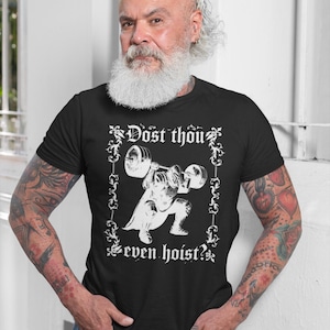Op de afbeelding: Zwart T-shirt met een witte afbeelding van een gewichtheffer en de tekst "Dost thou even hoist?" Het ontwerp is omlijst door decoratieve krullen. De persoon op de foto draagt het T-shirt en een blauwe spijkerbroek.