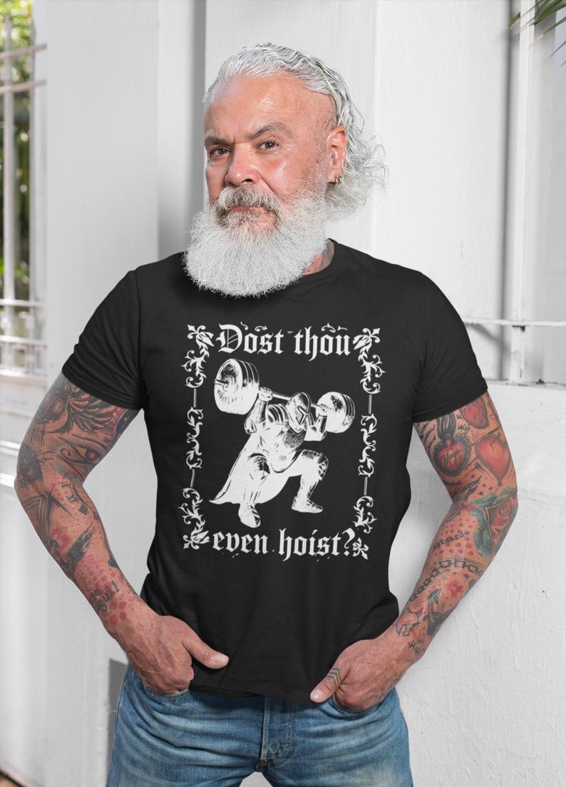 Op de afbeelding: Zwart T-shirt met een witte afbeelding van een gewichtheffer en de tekst "Dost thou even hoist?" Het ontwerp is omlijst door decoratieve krullen. De persoon op de foto draagt het T-shirt en een blauwe spijkerbroek.