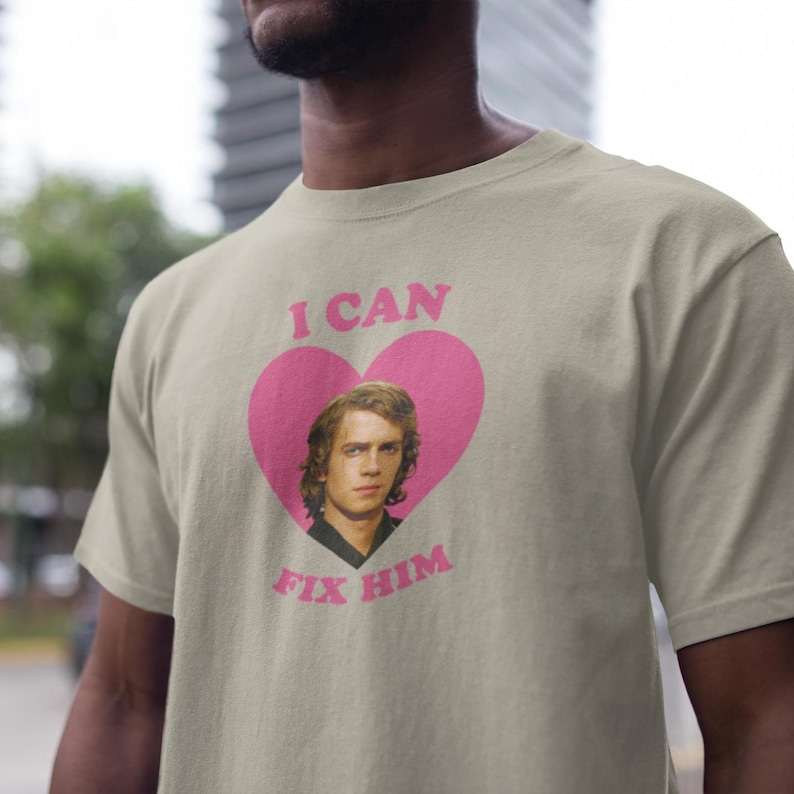 May include: A light beige t-shirt featuring a pink heart with the text "I CAN FIX HIM" and a portrait of a man. The text and heart are centered on the shirt.