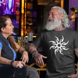 Puede incluir: Un hombre lleva una camiseta negra con un dise&ntilde;o de sol blanco. Una mujer est&aacute; sentada a su lado. El fondo incluye un bar con botellas y tarros.