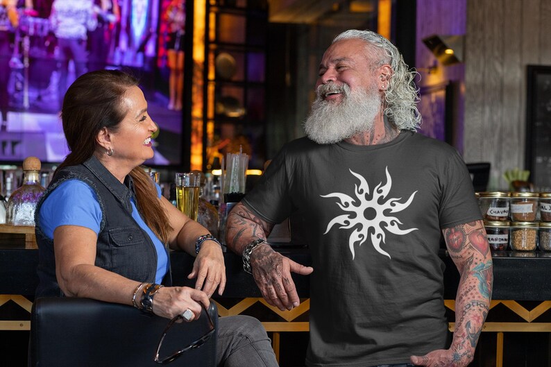 Puede incluir: Un hombre lleva una camiseta negra con un dise&ntilde;o de sol blanco. Una mujer est&aacute; sentada a su lado. El fondo incluye un bar con botellas y tarros.