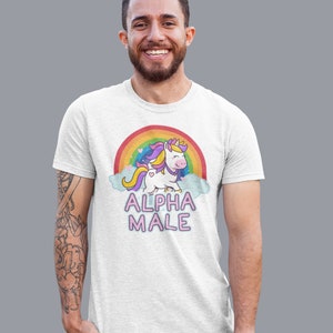 May include: White t-shirt featuring a cartoon unicorn with a rainbow and the text "ALPHA MALE". The unicorn has a yellow crown, purple and yellow mane, and a heart-shaped tail. The shirt is worn by a person.