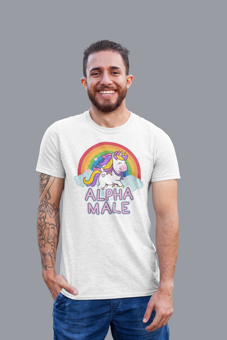 May include: White t-shirt featuring a cartoon unicorn with a rainbow and the text "ALPHA MALE". The unicorn has a yellow crown, purple and yellow mane, and a heart-shaped tail. The shirt is worn by a person.