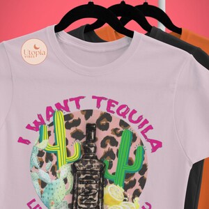 以下が含まれることがあります： ピンクのTシャツに、テキーラのボトル、サボテン2本、レモン、レオパード柄の円形のグラフィックデザインが施されています。円形の周りに「I want tequila」という文字がピンク色で書かれています。
