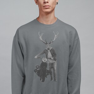 May include: Gray sweatshirt featuring a graphic of a deer-human hybrid figure with large antlers, wearing a jacket and jeans, leaning on a vintage motorcycle. The design is in grayscale, set against a gray background. The sweatshirt is a solid gray color.
