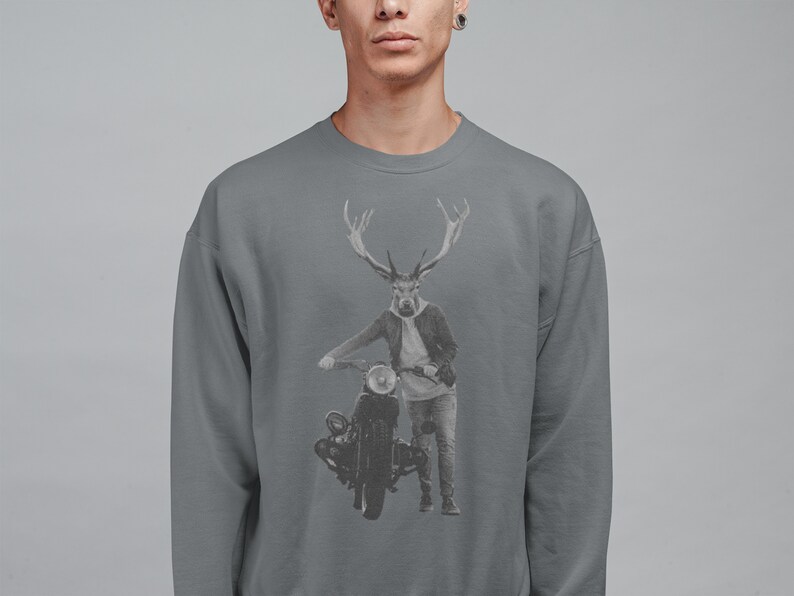 May include: Gray sweatshirt featuring a graphic of a deer-human hybrid figure with large antlers, wearing a jacket and jeans, leaning on a vintage motorcycle. The design is in grayscale, set against a gray background. The sweatshirt is a solid gray color.
