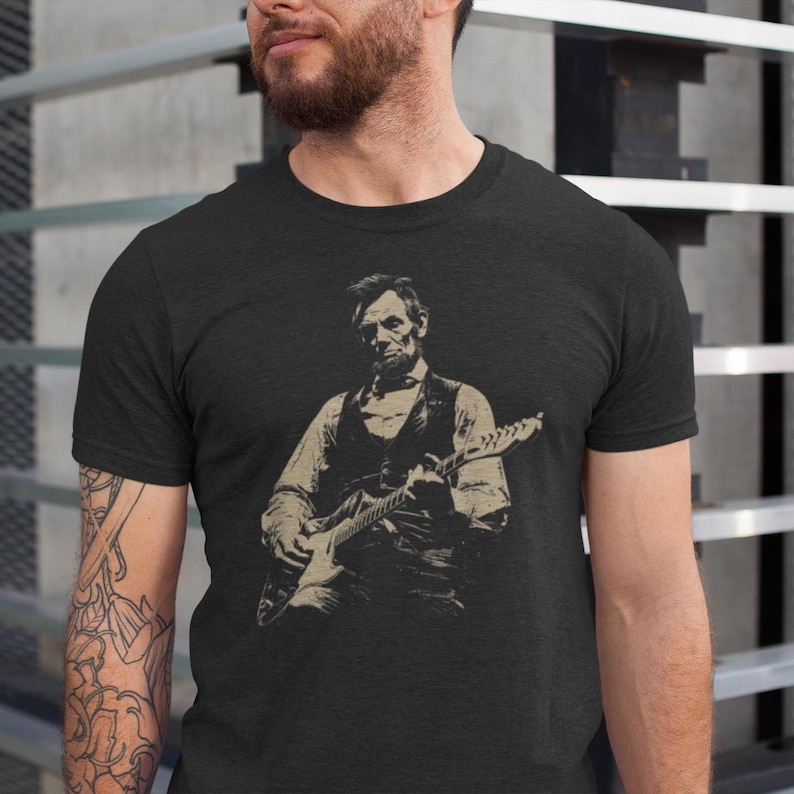 Peut inclure: T-shirt gris fonc&eacute; avec une illustration s&eacute;pia d'Abraham Lincoln jouant de la guitare &eacute;lectrique. L'image montre Lincoln en gilet et chemise, tenant la guitare. Le t-shirt a un col rond et des manches courtes.