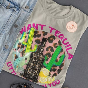 以下が含まれることがあります： 薄いグレーのTシャツに、ピンクと茶色のレオパード柄の円形がプリントされています。円形にはサボテン、テキーラのボトル、レモンが描かれています。円形の周りに「I want tequila, little bit of you」という文字がピンクで書かれています。