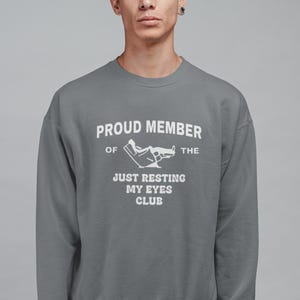 May include: A gray sweatshirt with the text "PROUD MEMBER OF THE JUST RESTING MY EYES CLUB" in white. The graphic includes a person reclining in a chair. The sweatshirt is a crew neck style.