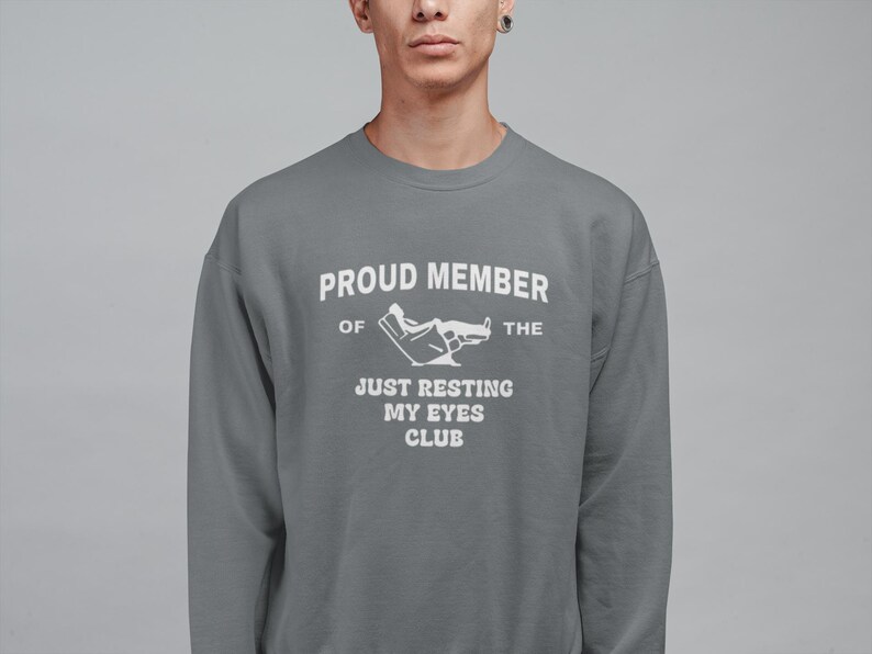 May include: A gray sweatshirt with the text "PROUD MEMBER OF THE JUST RESTING MY EYES CLUB" in white. The graphic includes a person reclining in a chair. The sweatshirt is a crew neck style.