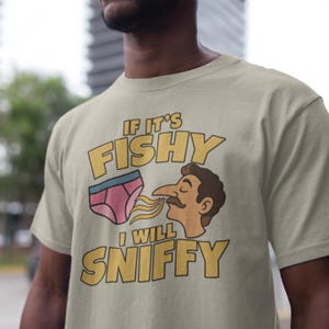 May include: A beige t-shirt featuring a graphic illustration of a pair of pink underwear and a person with a mustache sniffing them. The text reads IF IT'S FISHY I WILL SNIFFY in bold yellow letters with a black outline, centered on the chest.