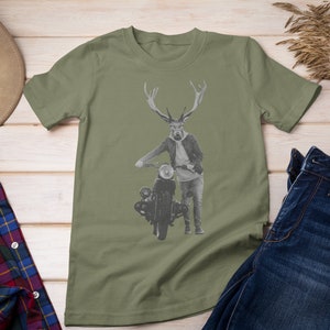May include: Olive green t-shirt featuring a deer-human hybrid with antlers, wearing a jacket and jeans, leaning on a vintage motorcycle. The image is set against a light wood background, with a hat and plaid shirt visible.