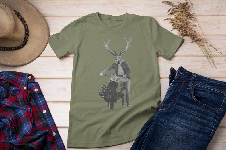 May include: Olive green t-shirt featuring a deer-human hybrid with antlers, wearing a jacket and jeans, leaning on a vintage motorcycle. The image is set against a light wood background, with a hat and plaid shirt visible.