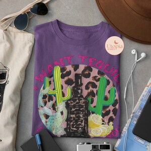 以下が含まれることがあります： ピンクとブラックのレオパード柄の円形デザインが施されたパープルのTシャツ。円にはピンク色の「I want tequila 4 u」の文字と緑色のサボテンのグラフィックが描かれています。円の真ん中にはテキーラのボトルがあります。