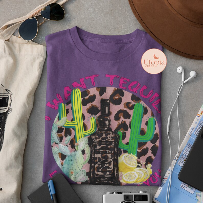以下が含まれることがあります： ピンクとブラックのレオパード柄の円形デザインが施されたパープルのTシャツ。円にはピンク色の「I want tequila 4 u」の文字と緑色のサボテンのグラフィックが描かれています。円の真ん中にはテキーラのボトルがあります。