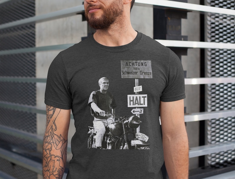May include: A dark heather grey t-shirt featuring a black and white photo of a man riding a motorcycle. The man is wearing a white t-shirt and jeans. The motorcycle is a vintage model with a sidecar. The t-shirt also features a sign that reads "ACHTUNG 100 m Schweizer Grenze 607 HALT".