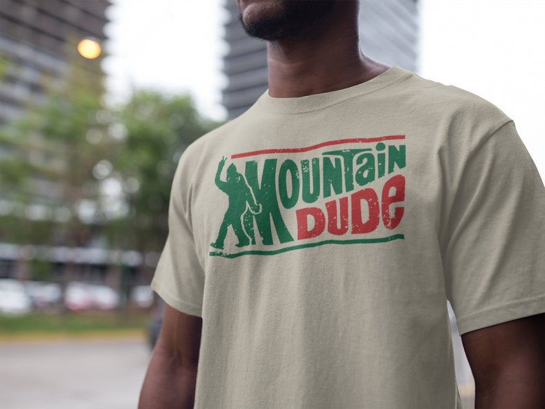 Może przedstawiać: Beżowa koszulka z zielonym i czerwonym nadrukiem graficznym z napisem "Mountain Dude". Grafika przedstawia sylwetkę stworzenia przypominającego Bigfoota.