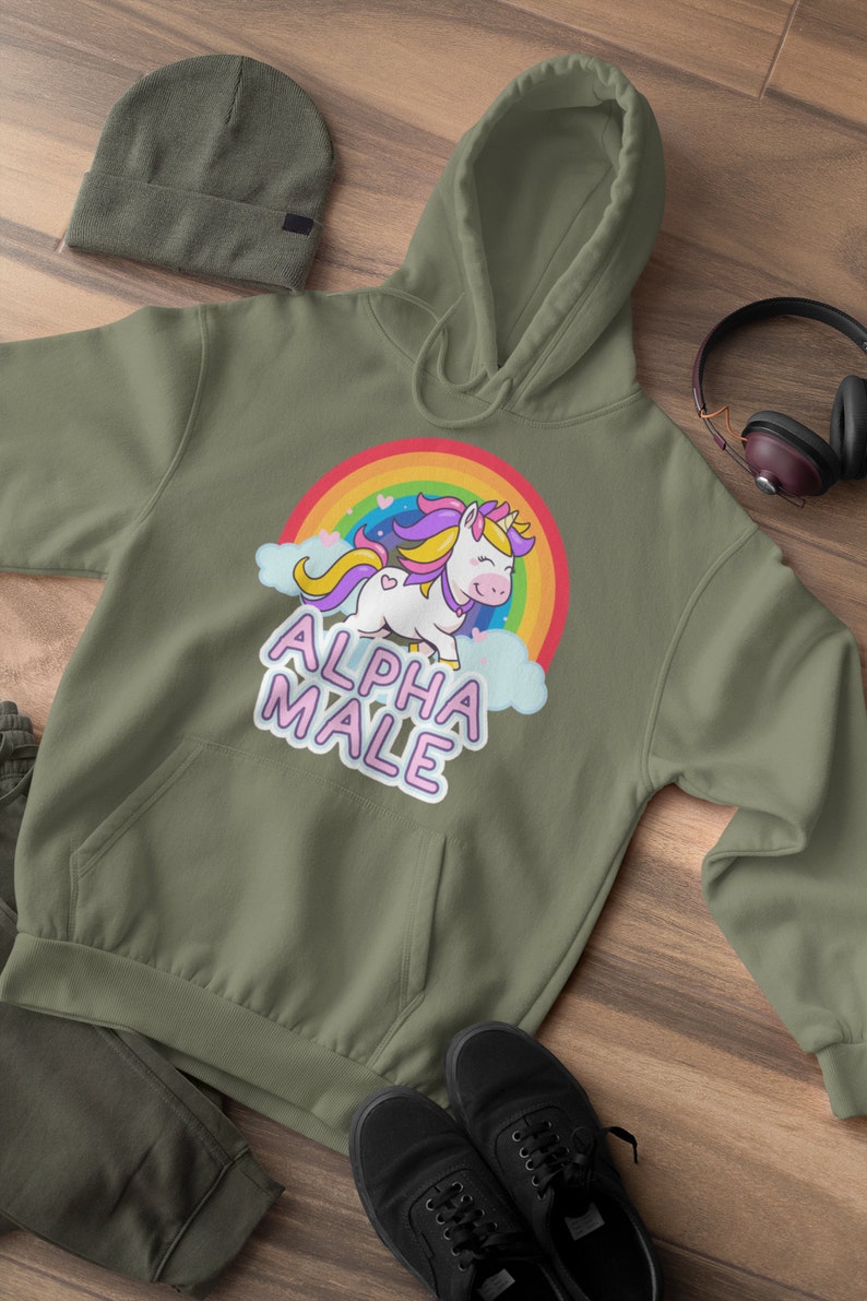 May include: Olive green hoodie with a front pocket, a drawstring hood, and a graphic of a unicorn with a rainbow and the text "ALPHA MALE". The image also shows a matching beanie, pants, and black sneakers.