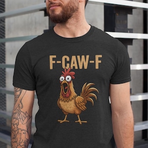 May include: Black t-shirt featuring a cartoon rooster with wide eyes and an open beak. The text "F-CAW-F" is printed above the rooster in a tan color. The shirt is a crew neck style.