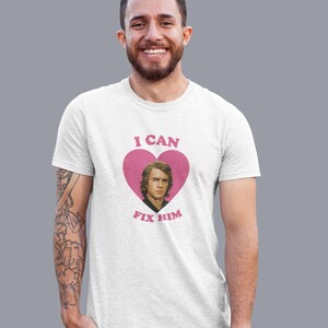 May include: White t-shirt with a pink heart graphic featuring a portrait and the text "I CAN FIX HIM". The shirt is worn by a person with a beard and tattoos on their arm.