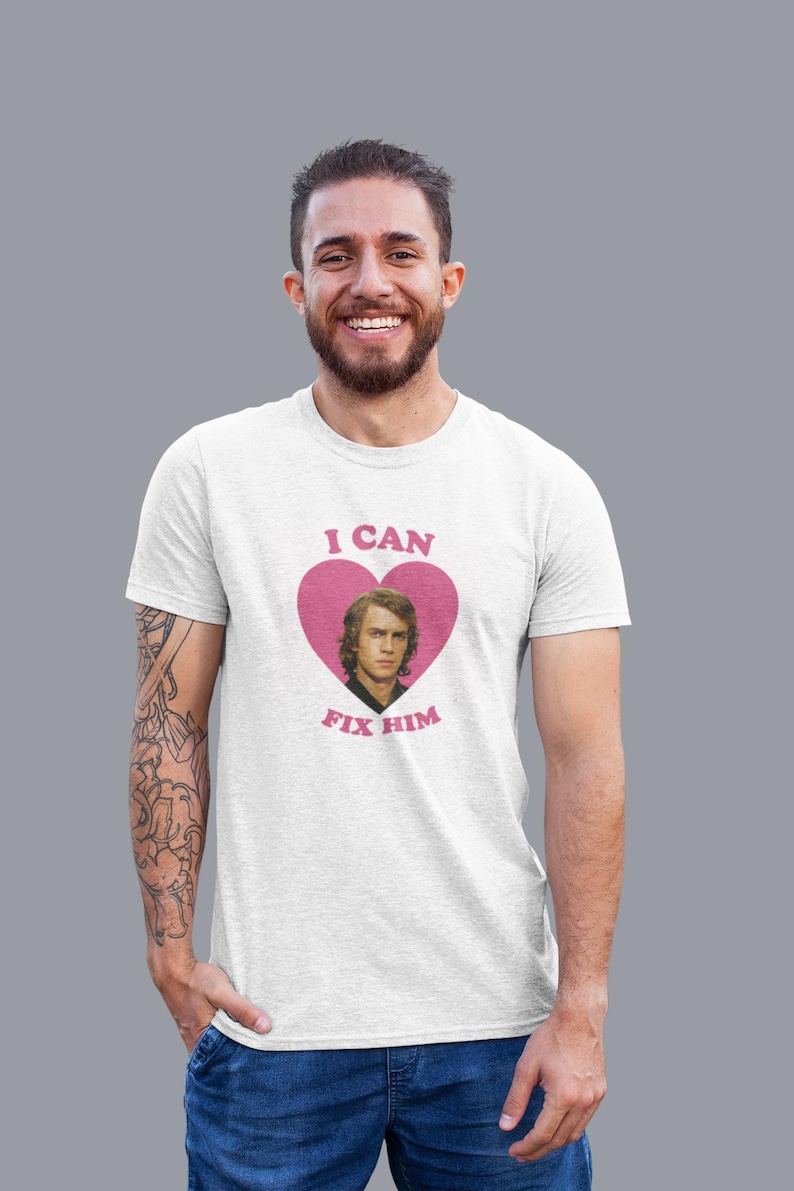 May include: White t-shirt with a pink heart graphic featuring a portrait and the text "I CAN FIX HIM". The shirt is worn by a person with a beard and tattoos on their arm.