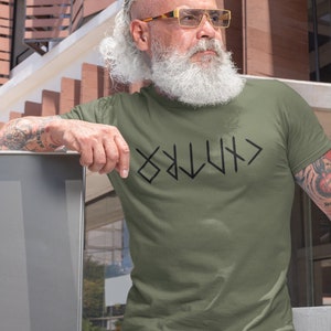 May include: A man wearing a green t-shirt with a black graphic of the word "VIKING" written in runes. He is wearing blue jeans and has a long white beard and gray hair. He is wearing gold-rimmed glasses.