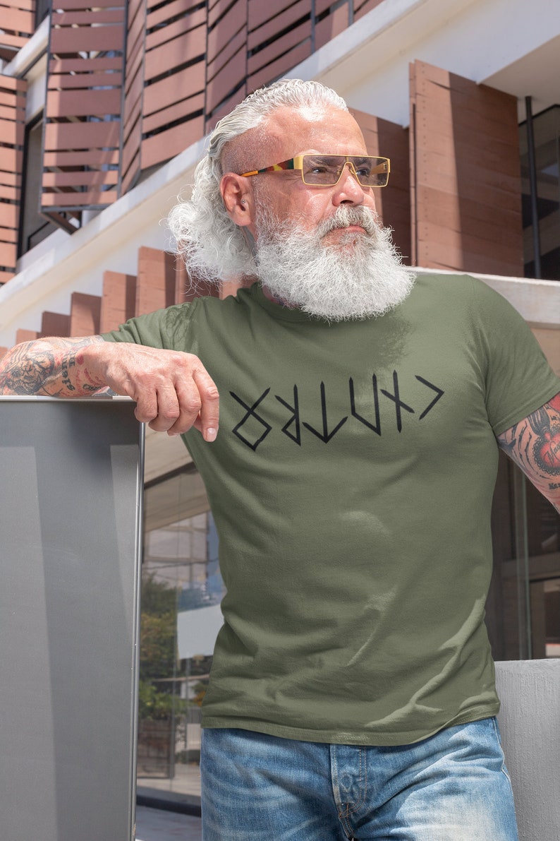 May include: A man wearing a green t-shirt with a black graphic of the word "VIKING" written in runes. He is wearing blue jeans and has a long white beard and gray hair. He is wearing gold-rimmed glasses.