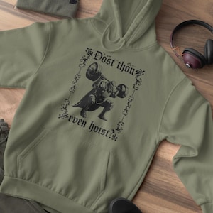 Op de afbeelding: Olijfgroene hoodie met een voorzak en een capuchon met trekkoord. De hoodie heeft een zwarte afbeelding van een gewichtheffer en de tekst "Dost thou even hoist?" Een bijpassende beanie, zwarte schoenen en een koptelefoon zijn ook zichtbaar.