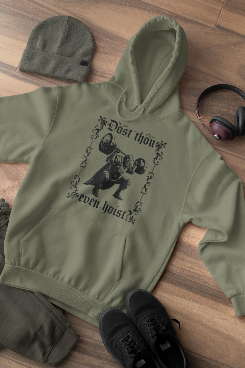 Op de afbeelding: Olijfgroene hoodie met een voorzak en een capuchon met trekkoord. De hoodie heeft een zwarte afbeelding van een gewichtheffer en de tekst "Dost thou even hoist?" Een bijpassende beanie, zwarte schoenen en een koptelefoon zijn ook zichtbaar.