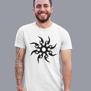 Puede incluir: Camiseta blanca con un dise&ntilde;o de sol tribal negro. El gr&aacute;fico del sol tiene un centro circular con formas de llama que se extienden hacia afuera. La camiseta est&aacute; hecha de una tela de color claro y la lleva una persona.