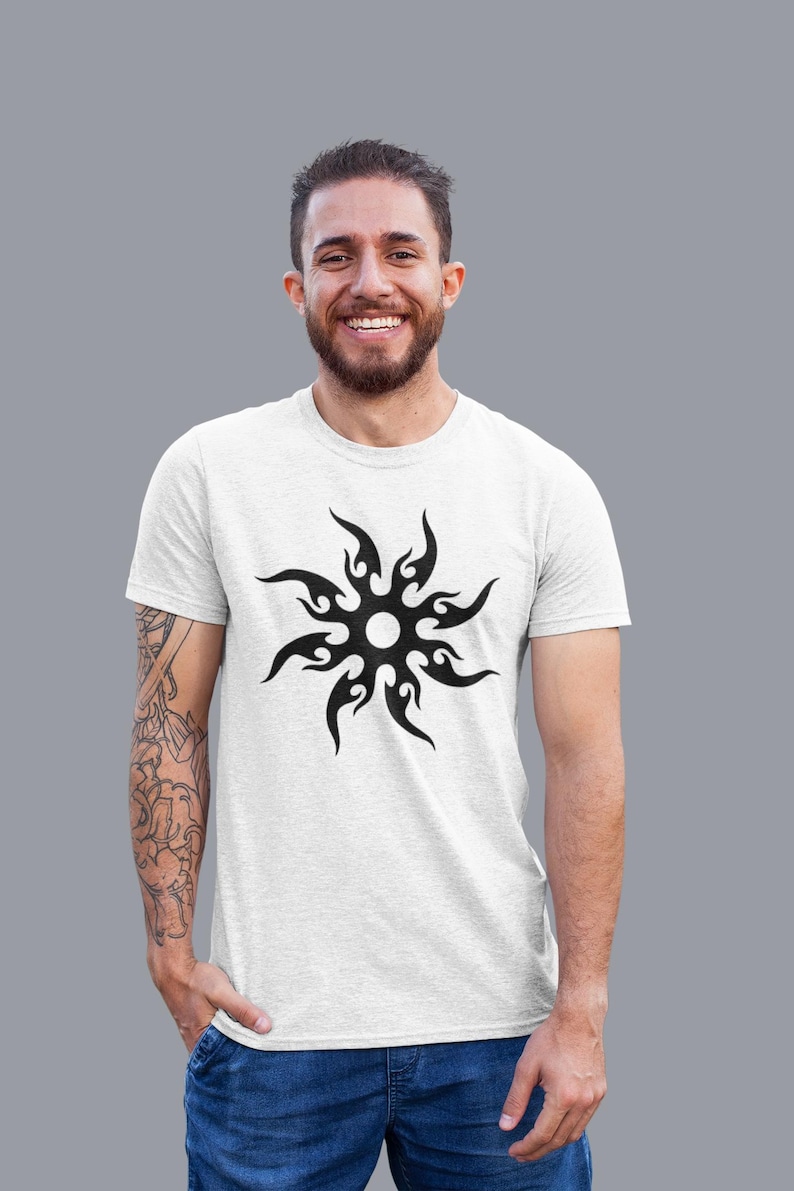 Puede incluir: Camiseta blanca con un dise&ntilde;o de sol tribal negro. El gr&aacute;fico del sol tiene un centro circular con formas de llama que se extienden hacia afuera. La camiseta est&aacute; hecha de una tela de color claro y la lleva una persona.