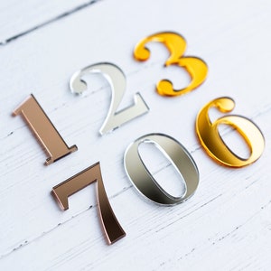 May include: A set of gold, silver, and rose gold numbers, 1, 2, 3, 0, 6, and 7, arranged on a white wooden surface.