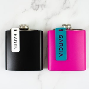 Op de afbeelding: Twee roestvrijstalen heupflacons, één zwart en één magenta, met zilveren doppen. De zwarte flacon heeft een wit label met de tekst "KAREN", en de magenta flacon heeft een turquoise label met de tekst "GARCIA".