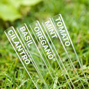 May include: Clear acrylic garden markers with white lettering. The markers are labeled with the names of herbs and vegetables: Cilantro, Basil, "Oregano", Mint, and Tomato. The markers are placed in green grass.