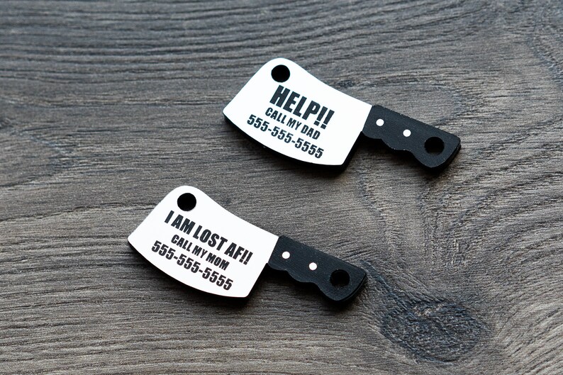 K&ouml;nnte beinhalten: Zwei wei&szlig;e Plastik-Hundemarken in Form eines Metzgermessers. Die obere Marke lautet "HELP!! CALL MY DAD 555-555-5555". Die untere Marke lautet "I AM LOST AF!! CALL MY MOM 555-555-5555".