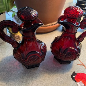 Può includere: Due caraffe di vetro rosso con disegni decorativi e tappi. Le caraffe hanno un motivo testurizzato, simile al diamante, e sono su una superficie di tessuto grigio chiaro.