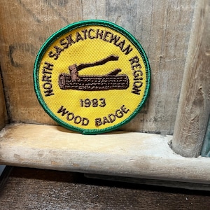 May include: A circular patch with a yellow background and green trim. The patch features the text "North Saskatchewan Region" and "Wood Badge" in black, with a brown log and axe graphic. The year "1983" is also displayed.